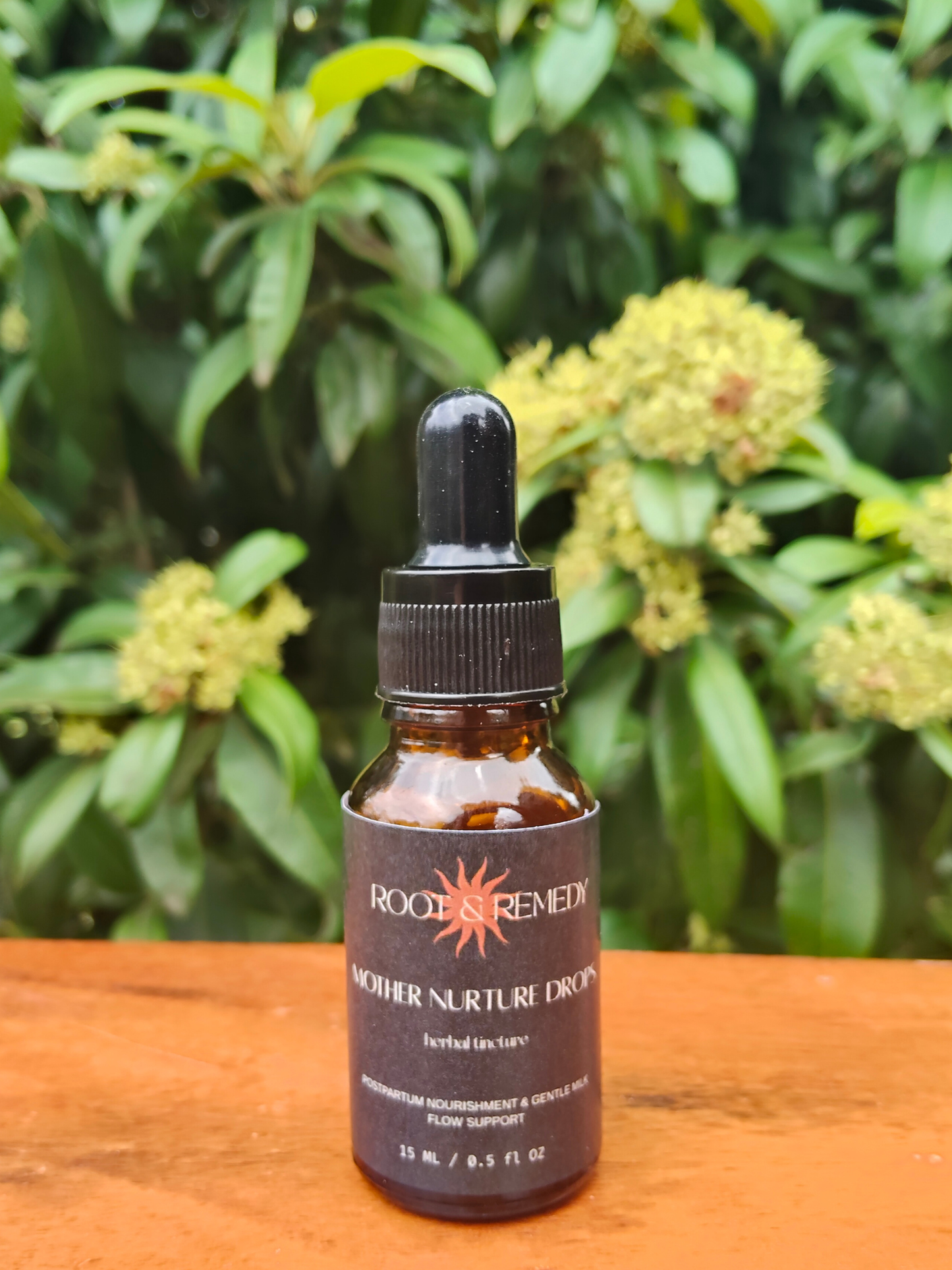 Bottle of 'Mother Nurture Drops' by Roots & Remedy on a wooden surface with green foliage in the background